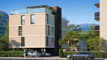 Modern multi-story building with a glass-enclosed terrace on the top floor, surrounded by greenery and mountains in the background.