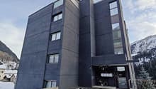 Modern multi-story building with multiple units, black exterior, several windows, entrance area, and a parking lot with a bicycle leaning against the building.