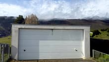 Garage with a white door, concrete walls, gravel driveway, fenced area, green field, mountains in the background