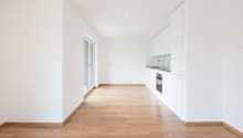 Empty room with wooden floor, white walls, large sliding glass doors, open kitchen, sink, oven.
