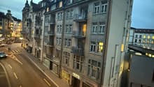 Urban apartment building, facade made of brick, glass windows, multiple floors, balconies, storefronts on the ground floor, traffic lanes, some parked vehicles, and street lamps on the sides.