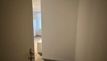 empty room, white walls, wooden floor, open door leading to bedroom, small white desk, white wardrobe, glass window with white curtain