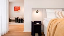 white bed with a blanket and pillows, dark night stand, lit lamp, white walls, picture on the wall, chair and table in the other room