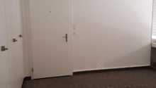 Small, rectangular room with two white doors, empty carpet floor, and white walls with visible lighting switches.