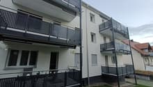 Modern apartment building with multiple balconies, white exterior, black railings, and a small garden in front.