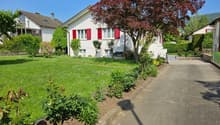 White house with red shutters, driveway, garden with various plants and a tree, neighboring houses in the background