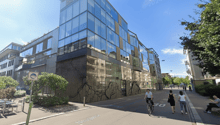 Modern building with glass facade, multiple floors, located on a street with people walking and a cyclist