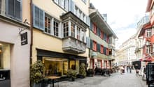 Cobblestone street, buildings with balconies, shops, people walking, outdoor seating, potted plants