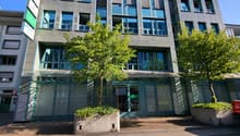 Multi-story office building with a modern, glass-and-concrete facade. The building has numerous windows, some of which appear to be balconies or terraces. There are trees and greenery surrounding the building, creating a pleasant, urban environment.