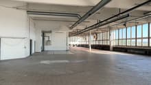 Large open commercial space with high ceilings, exposed ductwork, and tiled flooring. The space appears to be a warehouse or industrial facility with multiple windows along one side providing natural light. There are no visible furnishings or equipment, s