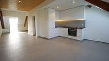 Modern, open-concept kitchen with white cabinets, built-in appliances including a dishwasher, and gray tile flooring. The kitchen is integrated into a larger living space with a vaulted ceiling and exposed wooden beams.