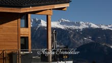 Wooden chalet-style building with a balcony overlooking snow-capped mountains in the background. The building has the name 'Angela Immobilier' displayed on the front.