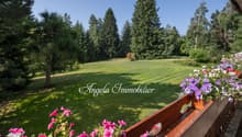 The image shows a scenic outdoor view from a balcony or terrace. The foreground features a wooden railing with colorful pink and purple flowers. Beyond the railing is a large, well-maintained lawn surrounded by tall pine trees. The sky is clear and blue,