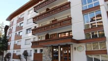 White and brown facade, 3 floors, large balconies with wooden railings, multiple windows, double doors at the entrance