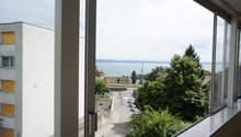 View from the window of a street with cars parked, a person walking, and a building with yellow shutters. In the distance, there is a body of water and a mountain range.
