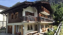 2-story wooden house, wooden balconies, white walls, wooden double doors, wooden picnic table, outdoor seating, wooden fence, grassy yard, mountainous landscape
