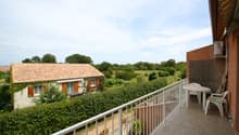 Two-story house with a tiled roof, surrounded by lush greenery and trees. The house has a balcony or terrace with a white railing, offering a scenic view of the surrounding countryside.