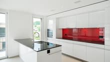 Modern, open-concept kitchen with white cabinets, red glass backsplash, and a large island with a black granite countertop. The kitchen features built-in appliances, including a dishwasher.
