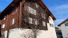 A building with a wooden wall, brick wall, and many windows. The house is surrounded by grass, and the snow is on the ground. It seems that the building is on the street. There is another building on the right, and it seems like there is a mountain in the