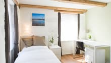 Cozy bedroom with exposed wooden beams, white walls, large window, bed with beige bedding, nightstand, and a desk with a chair.