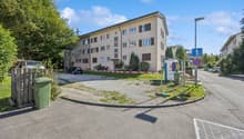 3 story building, beige exterior, multiple windows, cars parked in front, trees, street light, trash bin