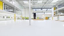 Large open commercial space with high ceilings, white walls, and yellow accents. The space has a stage-like area and appears to be a multipurpose event or exhibition hall.