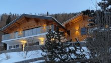 Two-story wooden chalet-style house with large windows, balcony, and snow-covered surroundings. The house has a warm, cozy appearance with exterior lighting illuminating the scene.