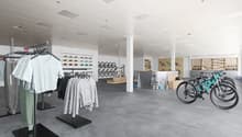 Large open space with clothing racks, shelves displaying accessories, and bicycles on display. The space has a modern, minimalist design with white walls, gray flooring, and recessed lighting.