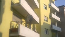 Yellow building with balconies, white awnings, glass doors, metal railings, metal satellite dish, garden with plants, outdoor table, chairs, and plants