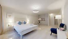Bed with white sheets and blue pillows, bedside tables with lamps, a blue chair, a kitchen with white cupboards, open floor design, two lamps and a table