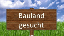The image shows a wooden sign in a grassy field, with the text 'Bauland gesucht' which means 'Land sought' or 'Land for sale' in German.