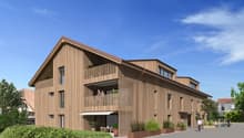 3-story wooden building with a slanted roof, balconies, and large windows. The building has a modern, eco-friendly design with a natural wood exterior. There are people visible on the balconies and a paved walkway in front of the building.