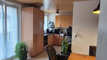 Dining area with wooden table, chairs, and a high chair. Wooden cabinets, kitchen appliances, and a window with curtains. Potted plants on the floor.