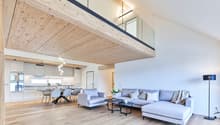 Spacious open-plan living area with wooden ceiling beams, large windows, modern kitchen with white cabinets, dining table with chairs, and a comfortable gray sofa set with coffee tables.