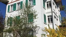 Two-story house with a traditional Swiss-style architecture, featuring a red-tiled roof, white walls, and green shutters. The house has a balcony or terrace on the upper level. The surrounding garden is lush with trees and shrubs, including a yellow flowe
