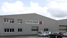 Large gray industrial building with the Alurex Kindt logo, multiple windows, and a parking lot in front of the building