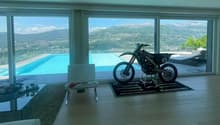 Large open-concept living room with floor-to-ceiling windows offering a panoramic view of the mountains and a swimming pool outside. The room features a modern design with a dirt bike displayed on a platform in the center.