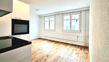 Empty room with wooden floor, white walls, two windows, built-in oven, and a sink.