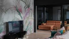 living room with large glass windows and balcony, wooden floor, teal sofa with cushions, coffee table, and wall mural, TV, and a rug