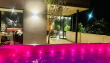 Jacuzzi with pink water and purple lights at night, next to an olive tree, with a dining area and a balcony behind