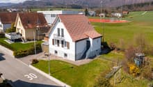 Detached house, brown roof, white walls, balcony, garage, garden, tennis court in the background