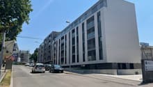 A modern multi-story commercial or residential building with a gray exterior and large windows. There is a parking area in front of the building with several cars parked.