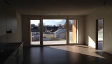 Spacious living room with large windows overlooking a balcony or terrace, dark tile flooring, and a view of the surrounding buildings and landscape outside.