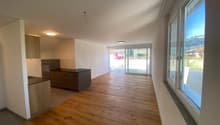 Open-plan living area with hardwood flooring, white walls, large windows providing natural light, and a sliding glass door leading to a balcony or terrace.