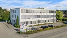 white office building with multiple windows, two stories, entrance, shrubs, parking area, bicycles parked under canopy