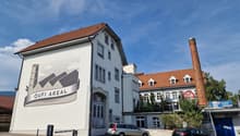 A multi-story building with a white exterior and a red tile roof. The building has several windows and a large sign on the side that says 'OUFI AREAL'. There are some cars parked in front of the building, and a tall chimney or smokestack can be seen in th