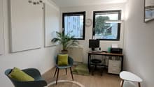 small room with wooden floor, white walls, two chairs, round rug, desk with computer, radiator, plant, clock, picture frame