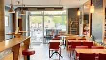a restaurant with a bar counter, several tables, chairs, red leather stools, lamps, menu boards, a framed picture on the wall, a blackboard, and a glass door leading to the outside