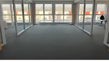 Large open office space with floor-to-ceiling windows providing a view of the surrounding buildings. The room has a gray carpet floor and a desk chair visible in the corner.