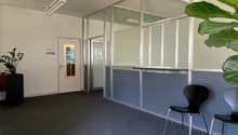 Empty office room with glass partitions, gray carpet, black chairs, a door, and potted plants